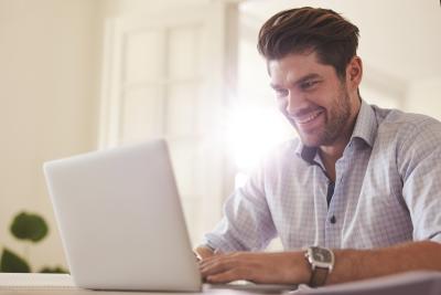 A man smiling at a laptop. Light is shining behind him.