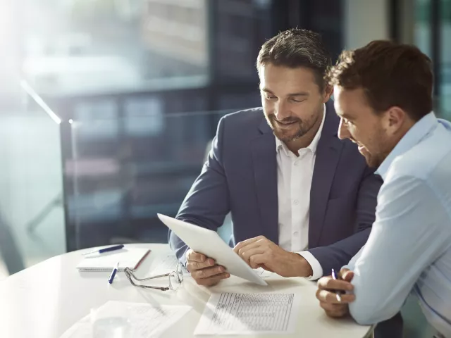 Two men sitting by the table and looking at a tablet. They're dressed in business attire.